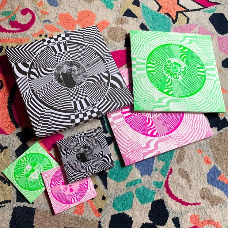 The album artwork for vinyl and CD showing the outer case in black and white 'optical illusion' pattern and the inner discs in sleees that have the same optical illusion pattern but instead in green and white instead of pink and white.