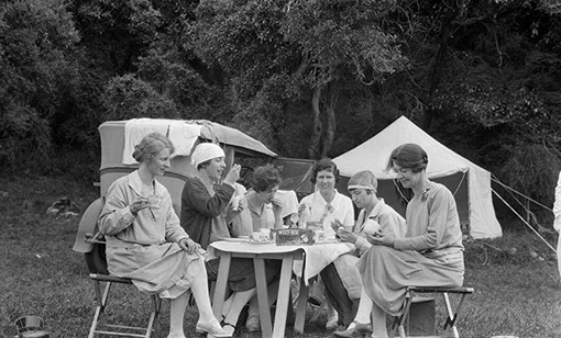 Group of six women having breakfast at a table while camping. There is a box of Weetbix on the table and some of the women are eating eggs.