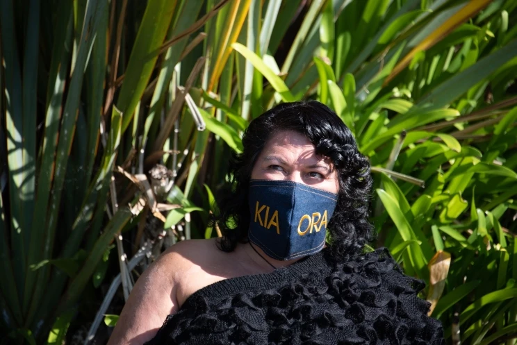 A woman standing with harakeke in the background and wearing a dark woven cloak and mask made of blue fabric with yellow words on the front.
