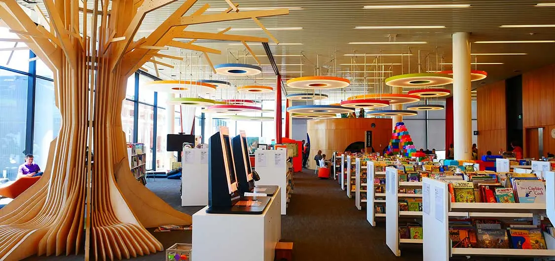 Photo of the children's area at Tūranga (Christchurch Central Library) with wooden tree-like sculpture, rows of shelving, toys, search screens, bright coloured ring-shaped ornaments hanging from ceiling.