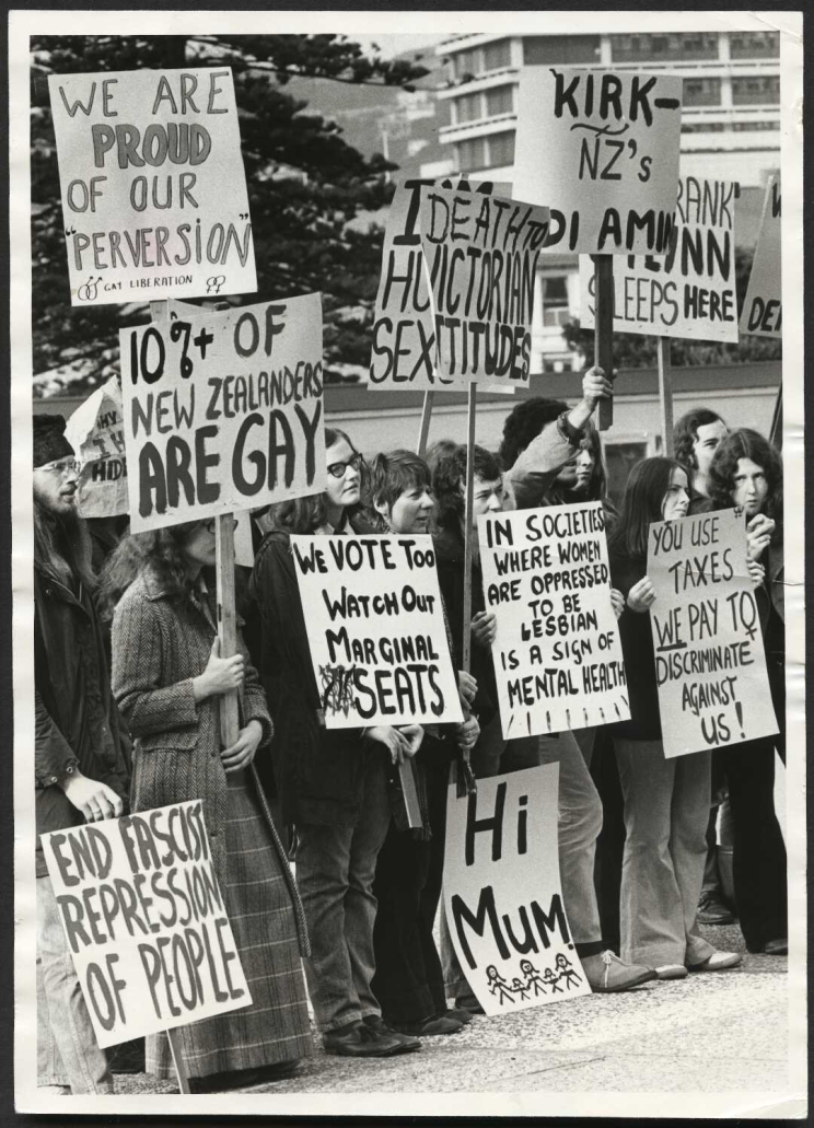 Black and white photo of picketers of all ages holding hand-made signs aloft outside Parliament. 