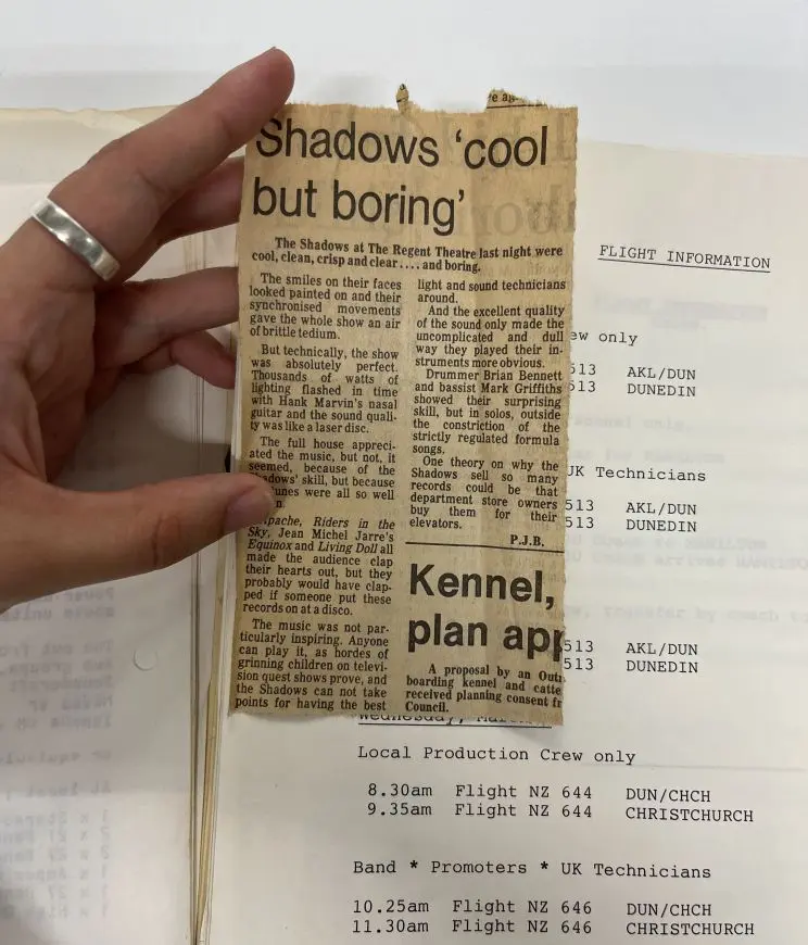 A hand holds a yellowed newspaper clipping showing the headline "Shadows 'cool but boring'" and behind this on a table is a folder with the arrival times of various technicians and crew from various destinations around New Zealand.