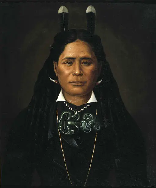 A striking portrait of a Māori woman. She Is wearing dark tailored clothing, has two huia feathers in her hair and a large tiki necklace. She is looking straight at us and has a Mona Lisaish smile. 