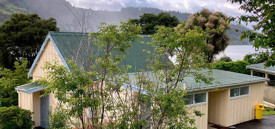 A school building surrounded by trees with water and tree-clad hills in the background.