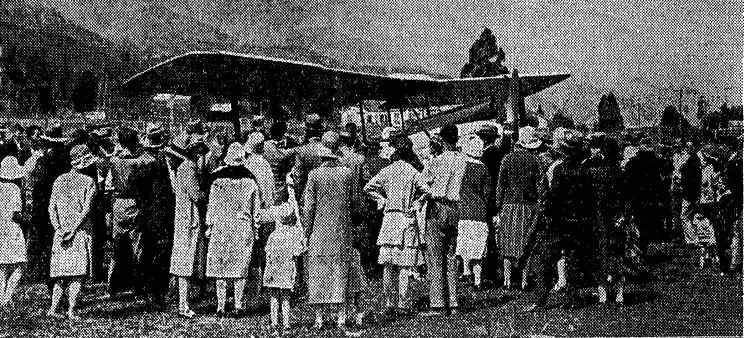 A grainy black and white image from a newspaper showing people standing around a biplane situated in a large field with hills in the background. 