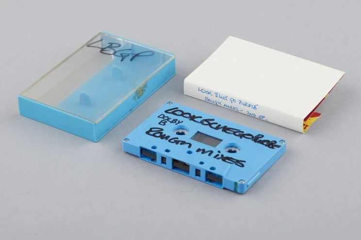 A view of blue cassette tape with black writing on outside alongside paper insert and plastic case with blue back.