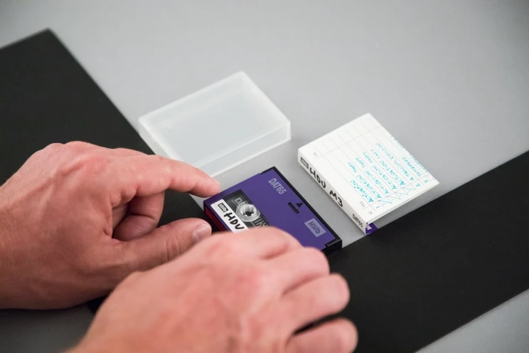 A small purple DAT tape with plastic cover and paper insert are being displayed neatly on grey hardboard in preparation for being photographed.