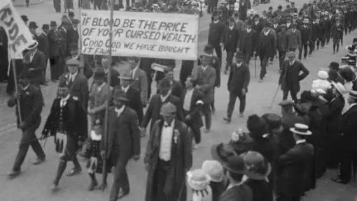 Shows strike action with men walking in a procession carrying large signs and banners. 