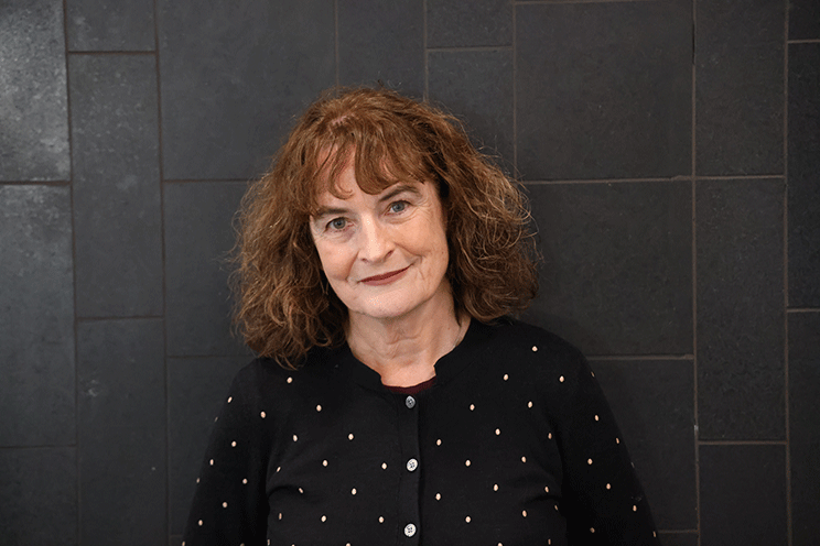 Photo of Kate De Goldi wearing a polka-dot cardigan, standing against a dark tiled wall.