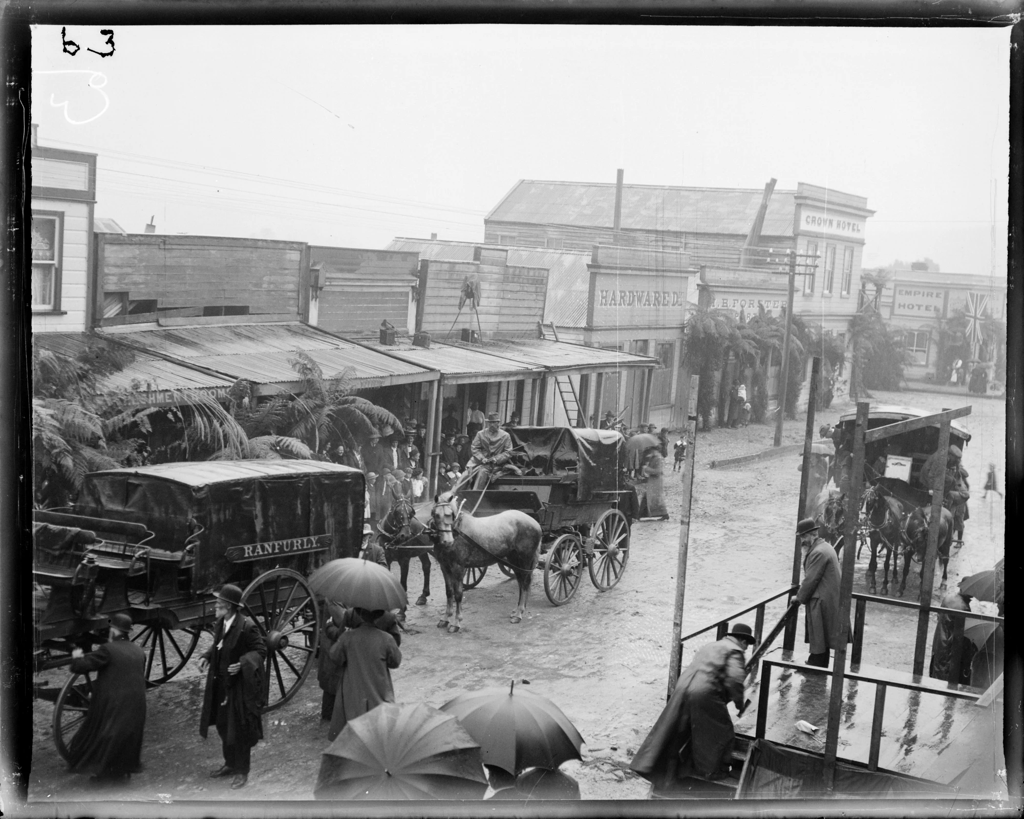 A rain-soaked street scene with horses and buggies waiting to pick up passengers and other pedestrians hurrying to shelter and carrying umbrellas.