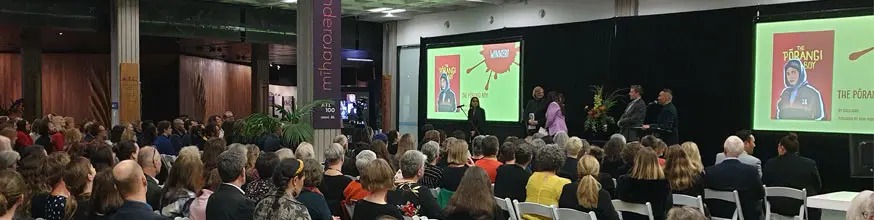 A large room of seated people looking at a stage with people on it and two digital screens with colourful images on them.