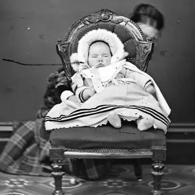 Victorian era black and white photo of a baby with a fur-edged hood and big dress sitting on an ornate dining room chair. A woman can be seen crouching behind the chair with one hand holding the baby. 