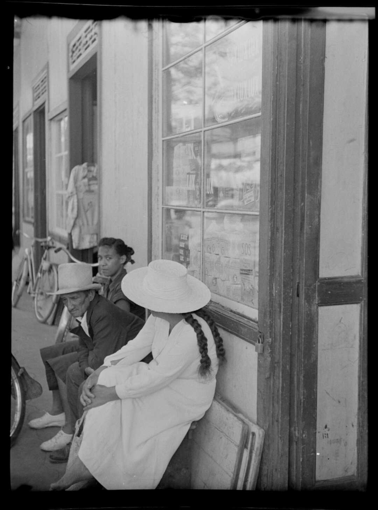 Three people sitting on a bench, two are wearing hats, a bicycle sits against the wall in the background.