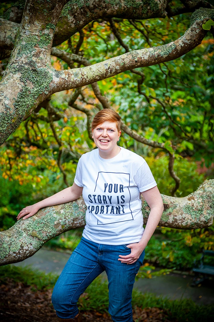 A woman wearing jeans and a white t-shirt with the words 'Your story is important', leaning against a tree branch.