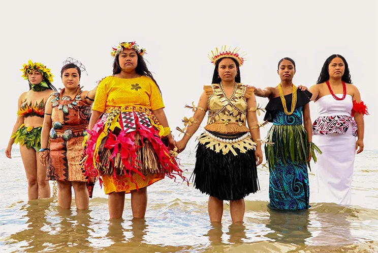 Six young women wearing traditional Pacific dress, standing in shallow sea water and looking directly at the camera.