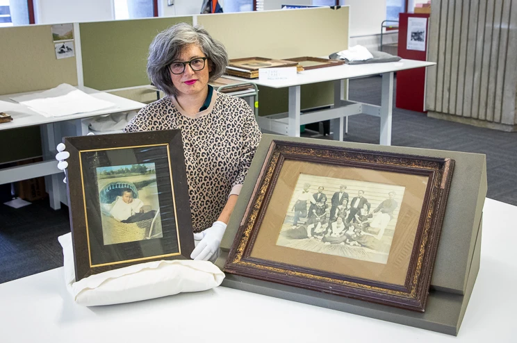 A woman wearing white gloves holds a framed portrait while another larger framed family portrait sits beside her on a table.