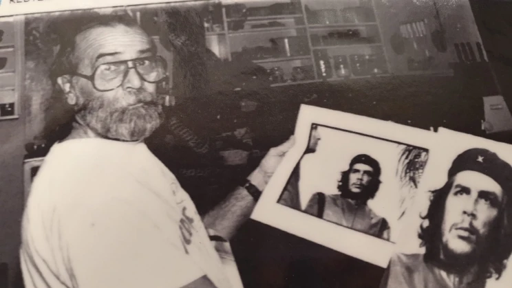 A black and white photo of a bearded man holding the iconic image of Che Guevara while looking back towards the camera.