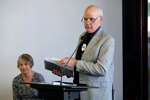 A man standing at a lecture holding a book, a woman sits near him.
