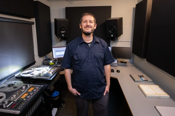 Portrait of AV conservator standing in his studio with tape machines and monitor speakers and other equipment.