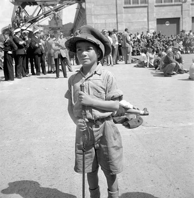 Black and white photograph of a young Māori boy standing in front of a crowd on the wharf. See Description below.