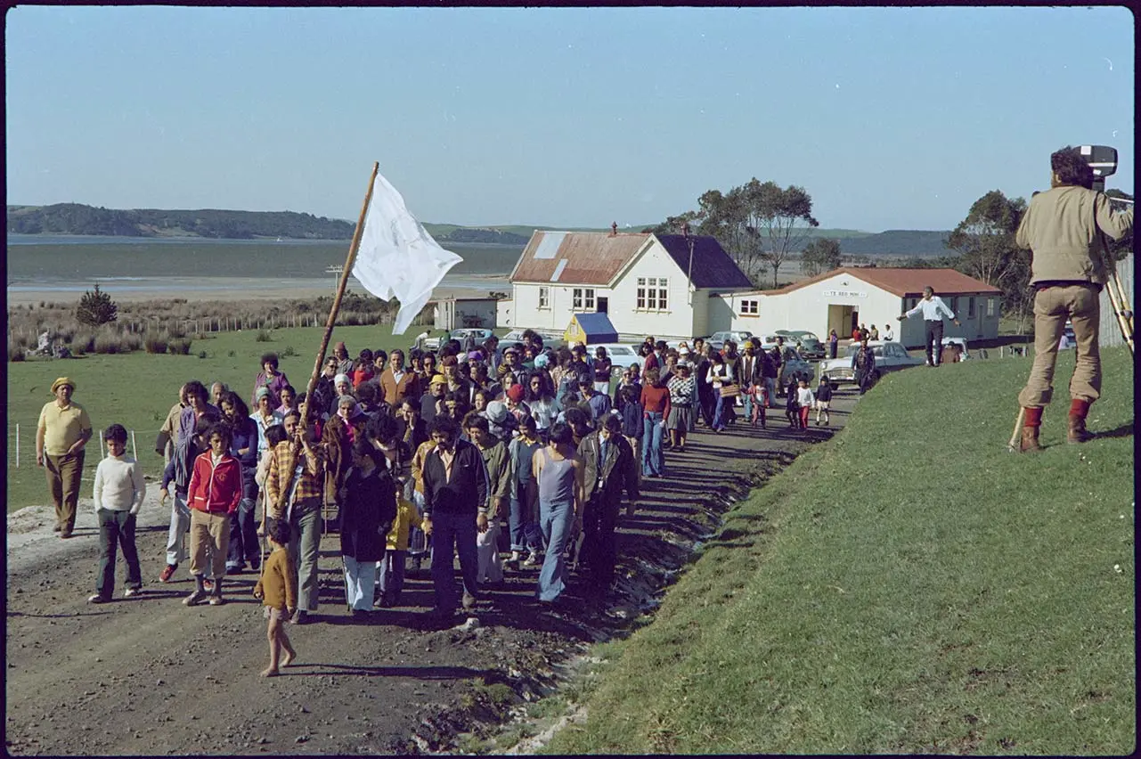 Colour photo of a hīkoi (march) — showing Māori Land March supporters leaving Te Hāpua. At the front, a Māori man carries a pouwhenua (land-marker post) with a white flag.