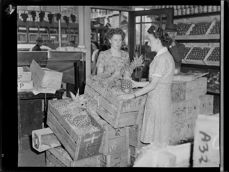 Women in a fruit shop looking at boxes of pineapples. 