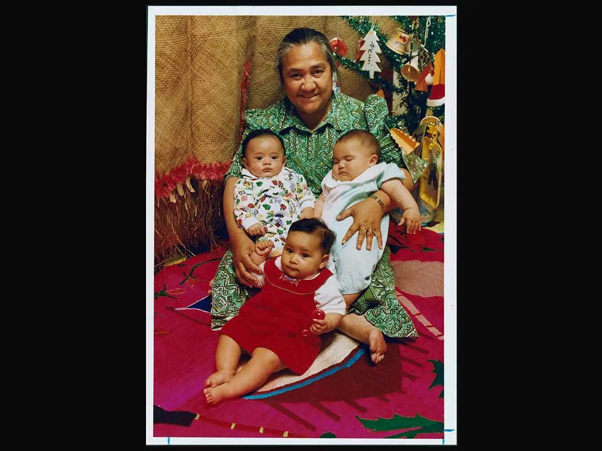 Fereni Ete holding 3 babies and sitting on a colourful mat. Behind them hang woven flax mats and a Christmas tree stands in the corner.