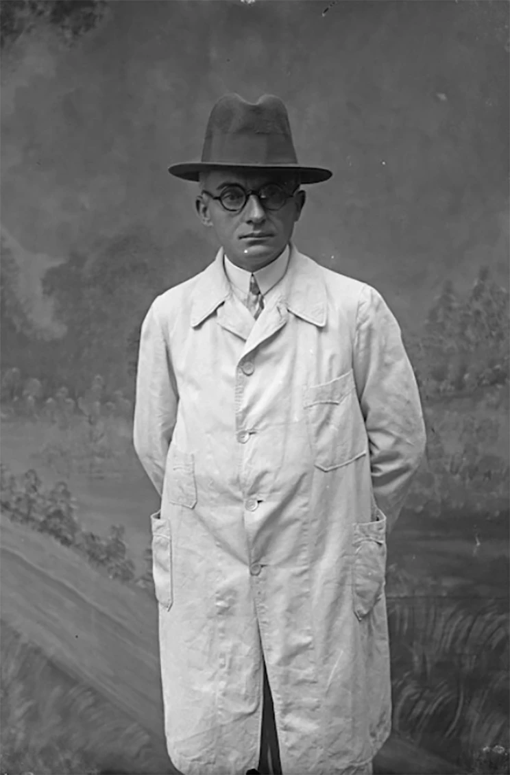 A man wearing an light coloured overcoat, a homburg hat and round glasses. 