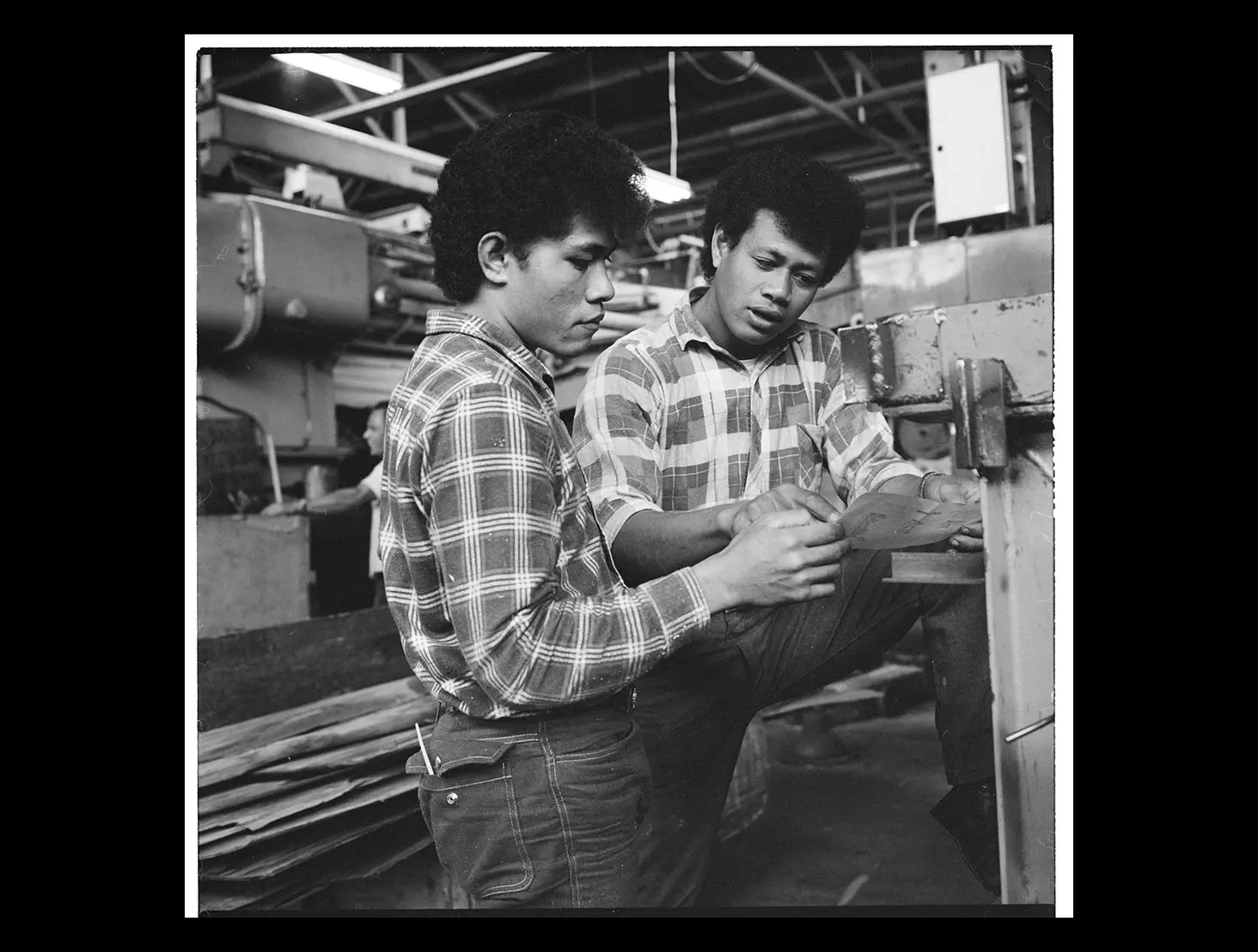 Black-and-white photo of 2 Pacific Island men working next to machinery at a plywood factory.