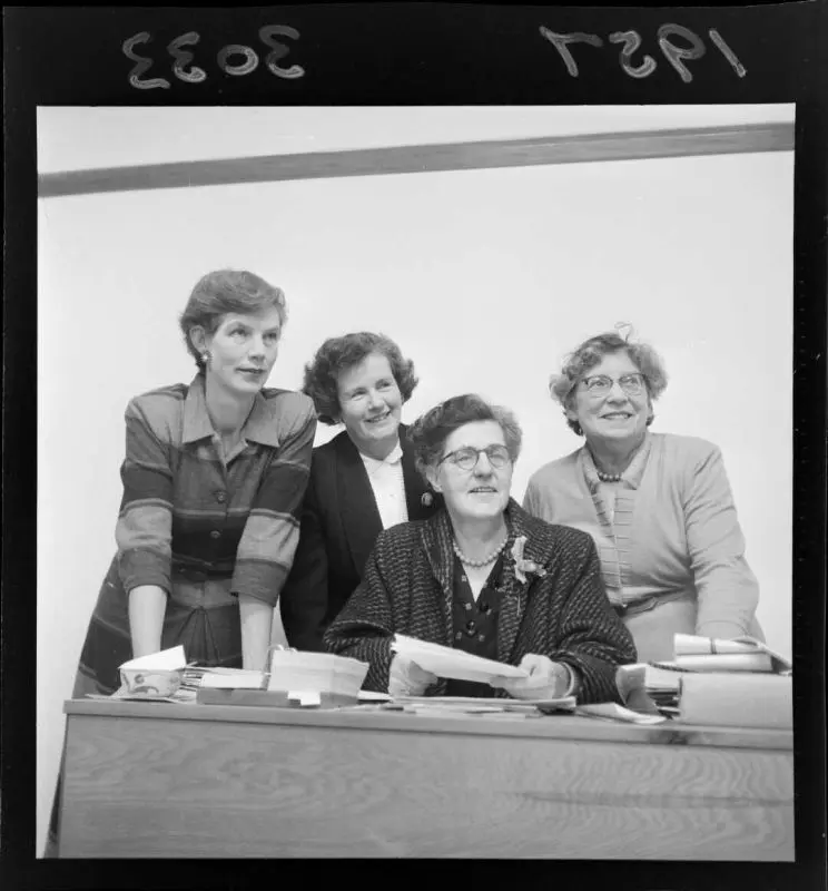 A black and white photo of four women behind a desk, looking off-camera and smiling. 
