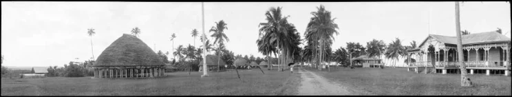 Panoramic view of a village in Samoa (probably near Apia) a person holding an umbrella walks towards the camera down an avenue of tall palm trees, on the left a house with open verandahs.