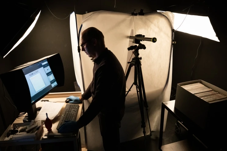 Imaging technician stands in the studio with camera, tripod and light tent in background, using specially calibrated computer monitor to check and modify images.