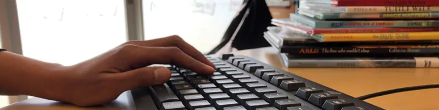 Hand on a keyboard with a pile of books in the background. 