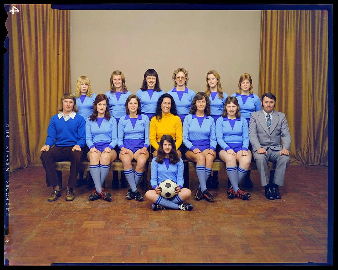 A colour team portrait showing two rows of women wearing their jerseys along with the captain in the middle in yellow, two male coaches on either side and a woman seated on the ground in front holding a soccer ball. 