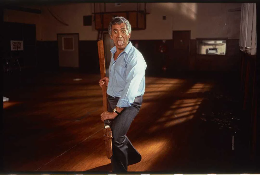 Man facing the viewer in a challenging side stance. The man is holding taiaha and performing a pūkana.