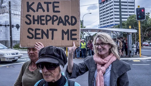 Three women on a street, two are holding a cardboard sign reading 'Kate Sheppard sent me'