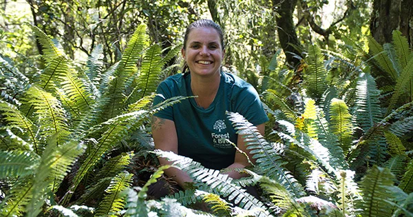 A smiling woman wearing a green t-shirt with the Forest & Bird logo crouching among ferns in the bush.