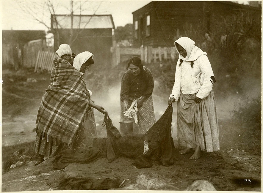 Black and white photograph of 3 Māori women holding edges of cooking cloths or a billy and bending over a steam vent. See Description below.