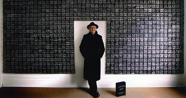 A man wearing a black hat and coat standing in front of an artwork.