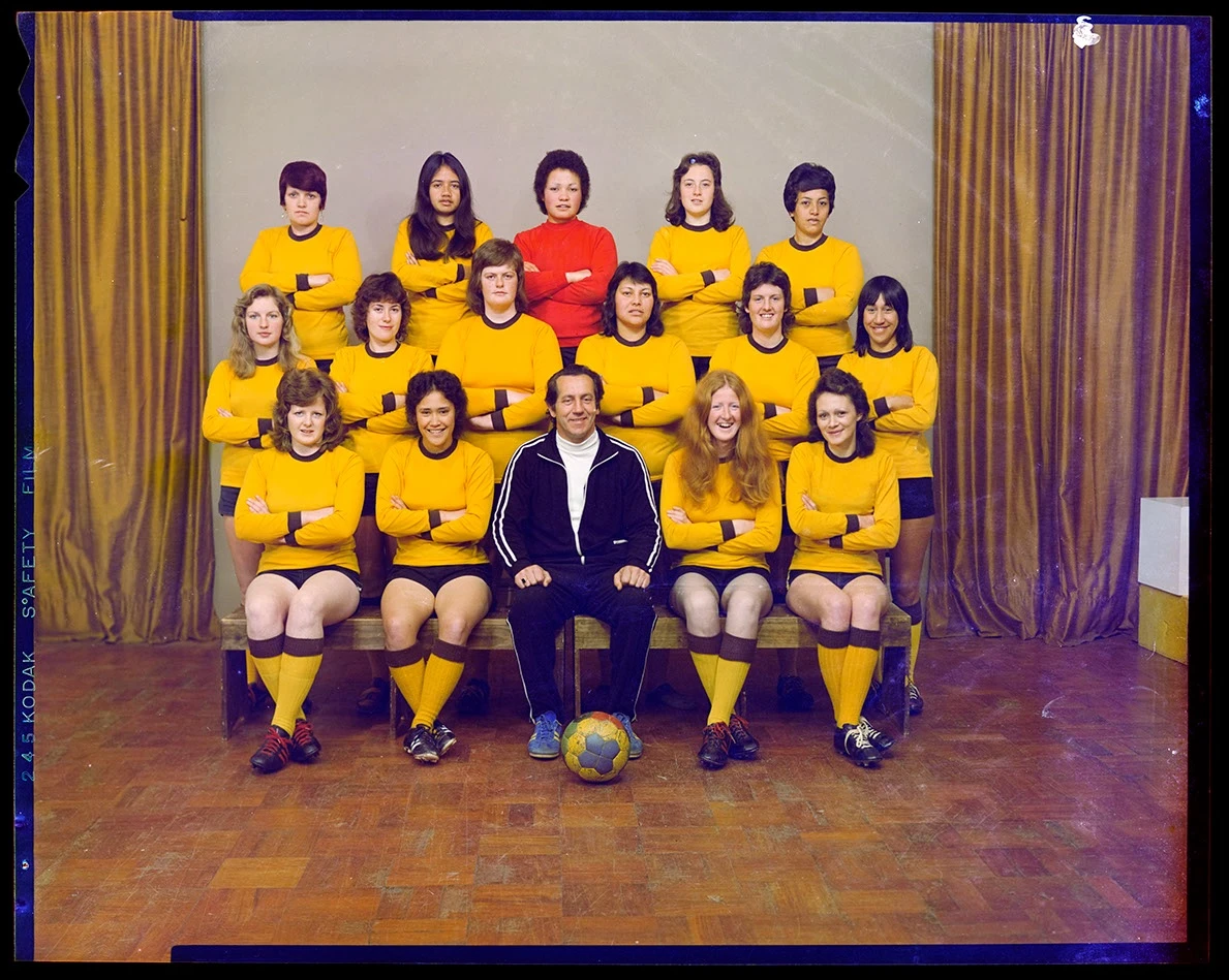 A colour studio portrait of a women's soccer team wearing yellow uniforms with the captain in red jersey in the middle of the top row and the male coach seated in the middle of the bottom of three rows with a football at his feet. 