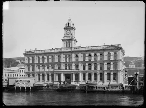Black and white photo of a large, three-storey building on a wharf. The building has many arch windows and an ornate clock tower. 