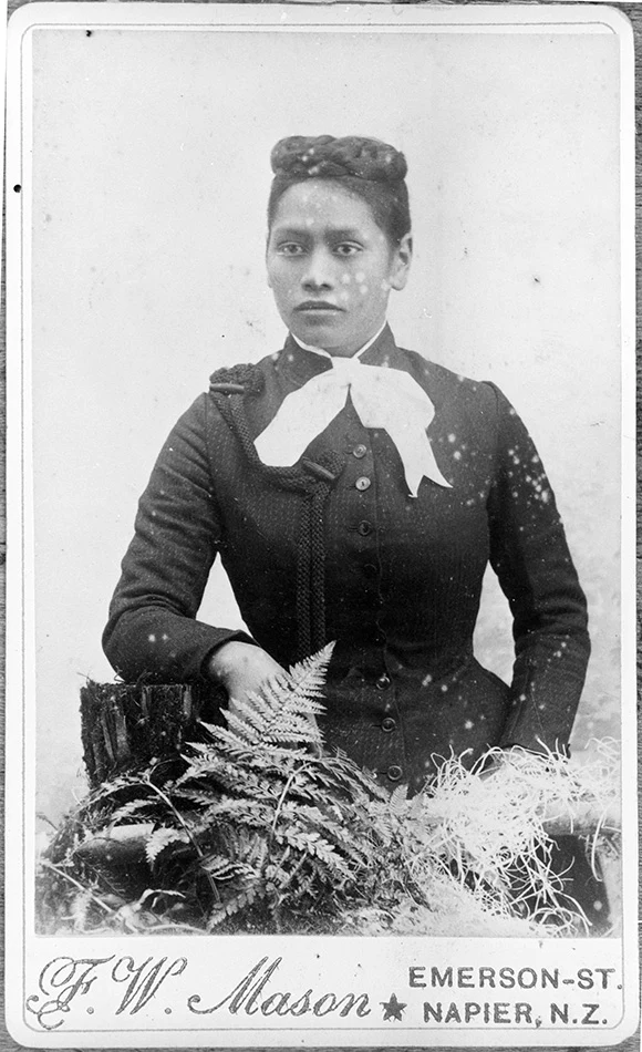 Copy of a cabinet card with a black and white studio photograph of Meri Te Tai Mangakāhia seated with plants. Below the photo are the words 'F W Mason Emerson Street Napier NZ'.