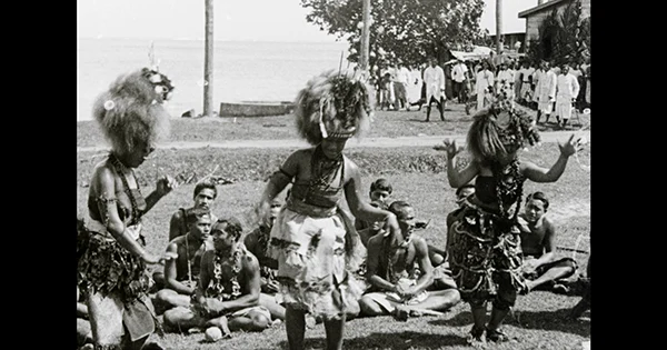 Three Samoan women performing a traditional dance with a crowd of people on the grass behind.