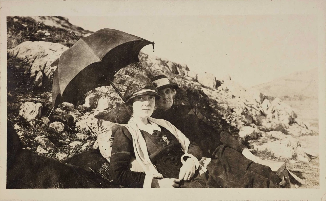 Katherine Mansfield reclining beside rocks with a friend, both under an umbrella to keep out of the sun.