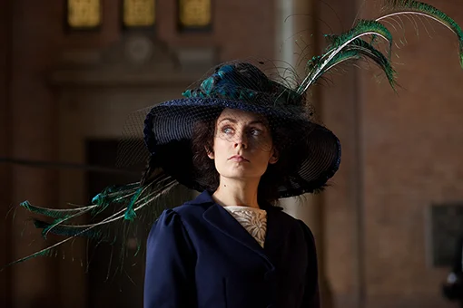 Actress Kate Elliott wearing an Edwardian style navy hat and jacket with peacock feathers.