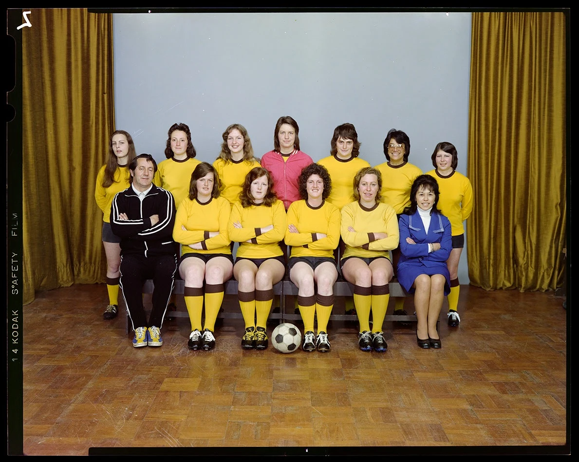 A studio portrait of a women's soccer team with the players wearing yellow uniforms, the captain in the middle of the top row wearing a red jersey and a male and female coach seated at either end of the bottom row. 