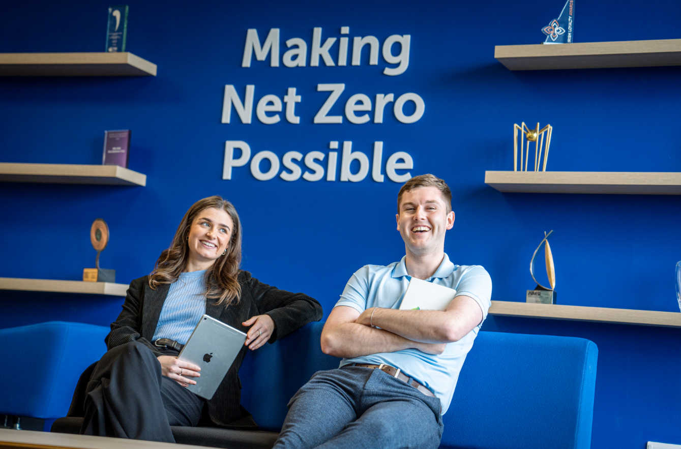 Two people smiling in office with the words 'Making Net Zero Possible' on a blue wall behind them. 