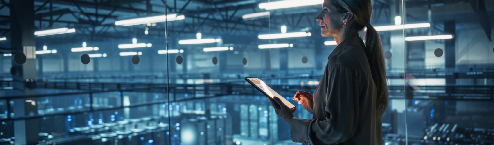 A woman in a suit, holding a tablet in the centre of what appears to be high-tech, digital equipment, resembling an interior of a data centre.