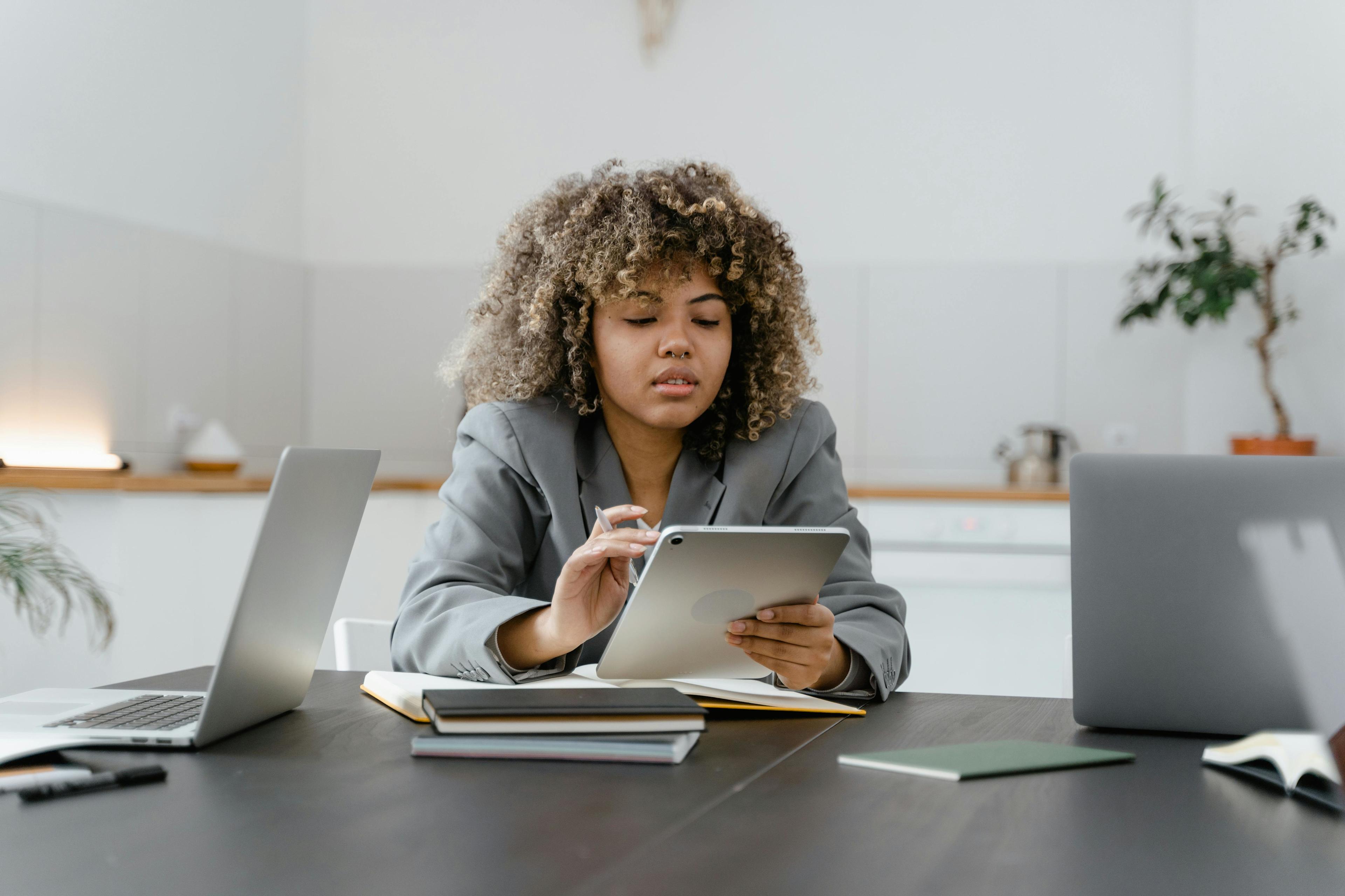 A lady sat at a table, checking her tablet, amongst some files and an open laptop.