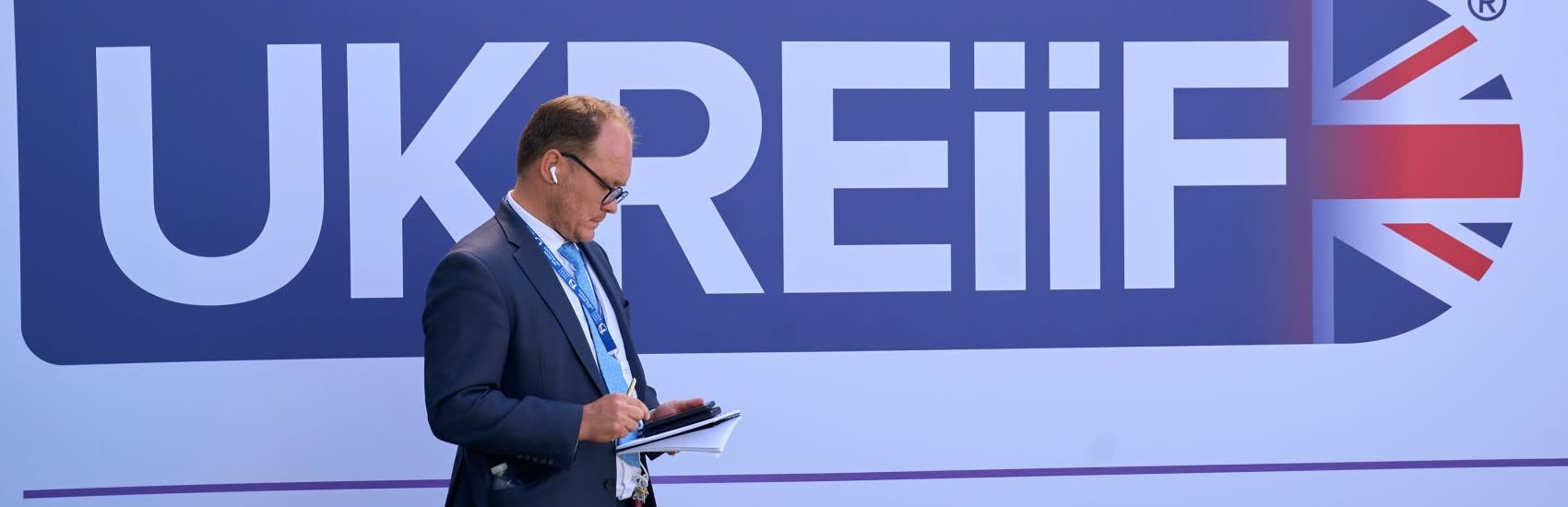 A UKREiiF logo in the background with a man in a blue suit standing in front of it, looking at his phone.